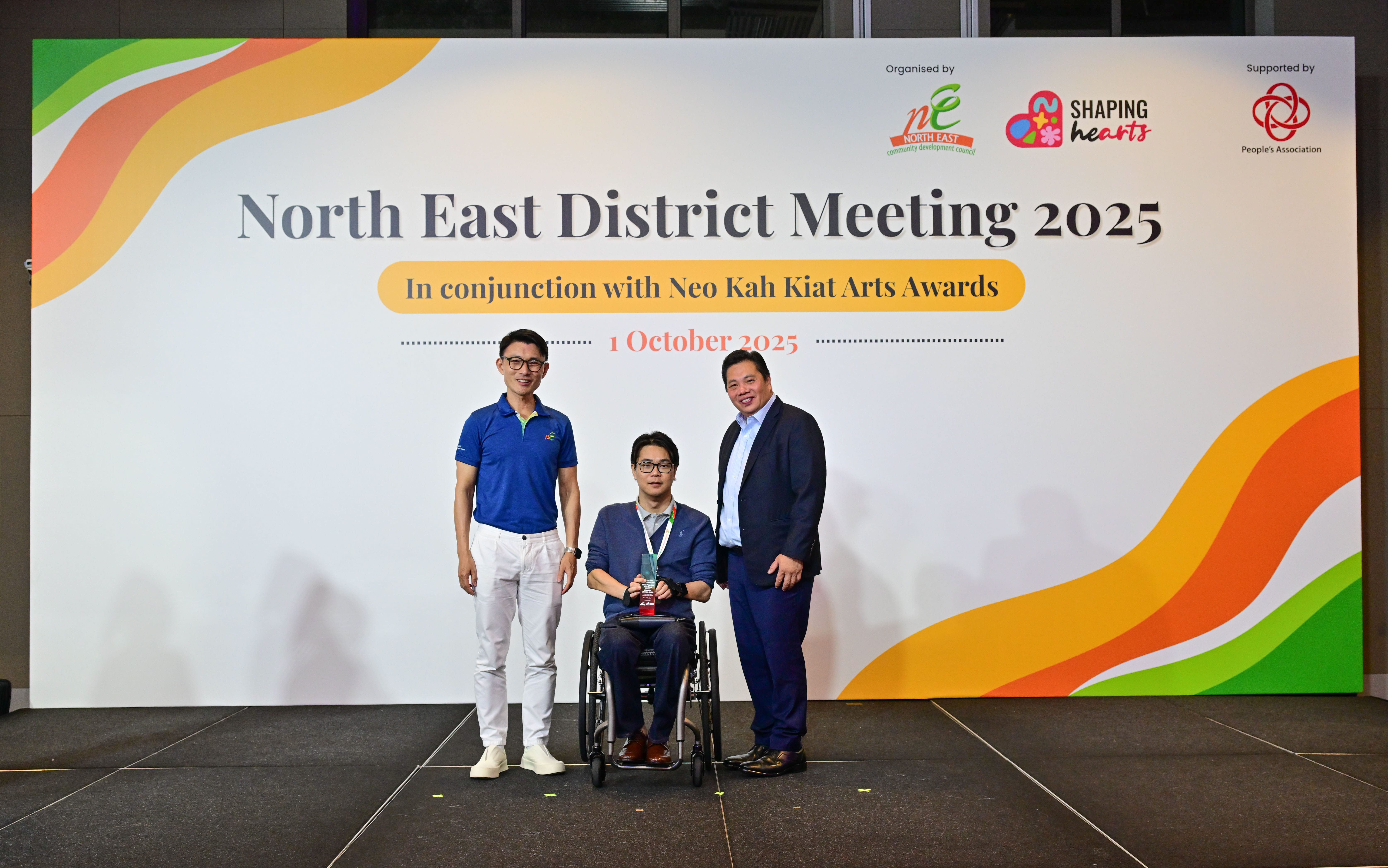 Award recipient standing on stage with the presenter, holding a acrylic plaque and posing for a group photo during the award ceremony, with a backdrop reading ‘North East District Meeting 2025 In conjunction with Neo Kah Kiat Arts Awards'